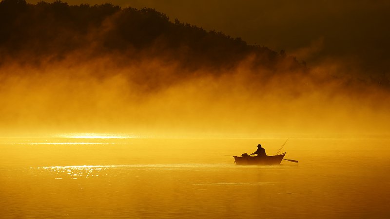 Bei Sonnenaufgang ist ein Angler in seinem Ruderboot mitten auf einem See zu sehen. Der See liegt in goldenem Licht.