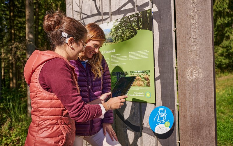 Eine Frau sowie eine Frau mit einem Cochlea-Implantat stehen vor einer Informationstafel im Nationalpark-Hunsrück-Hochwald und scannen mit ihrem Tablet einen QR-Code.