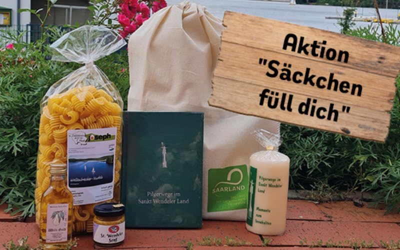 Für ihre nachhaltige Anreise erhalten Gäste ein Dankeschön in Form von Nudeln, einem Beutel, einer Kerze, einem Pilgerbuch, einem kleinen Getränk sowie Senf. Ein illustriertes Holzschild mit der schwarzen Aufschrift "Aktion Säckchen füll dich" steht darauf.