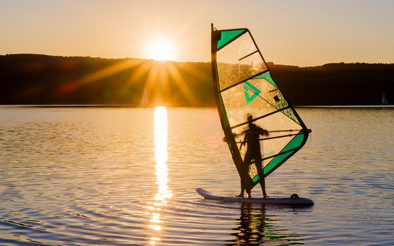 Im Hintergrund geht die Sonne hinter den Hügeln am Ufer unter. Ein Windsurfer befindet sich auf dem Bostalsee. Er ist im Vordergrund zu sehen.