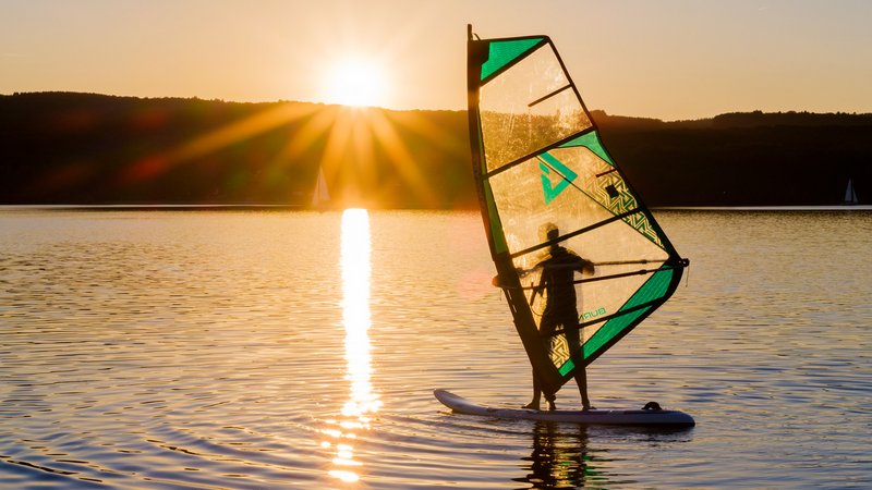 Im Hintergrund geht die Sonne hinter den Hügeln am Ufer unter. Ein Windsurfer befindet sich auf dem Bostalsee. Er ist im Vordergrund zu sehen.