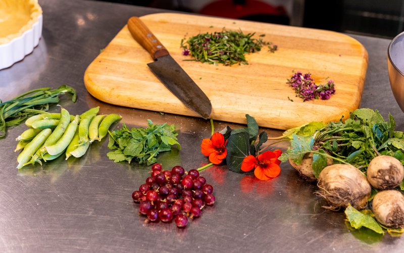 Auf einem Holzschneidebrett liegt ein Messer zum Hacken von Kräutern. Davor befinden sich verschiedenes Gemüse, Beeren und Kräuter.