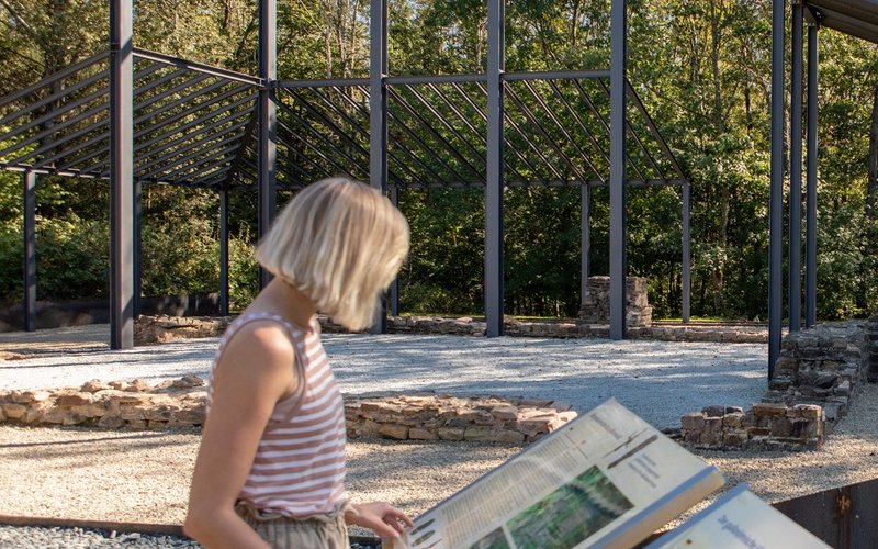 Eine Frau studiert eine Objekttafel des Vicus Wareswald. Im Hintergrund sind Kunstbauwerke zu sehen.