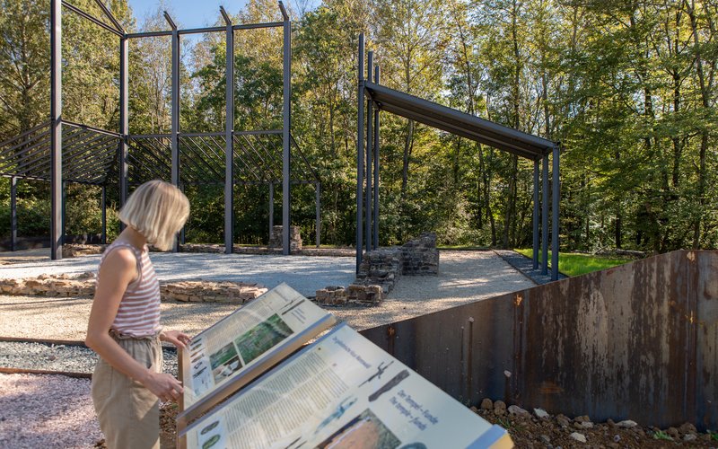 Eine Frau studiert eine Objekttafel des Vicus Wareswald. Im Hintergrund sind Kunstbauwerke zu sehen.