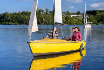 Ein gelbes Segelboot im Vordergrund mit einer Frau und zwei Männern als Insassen steuert schräg auf die Kamera zu. Die Frau, mit den Seilen des Segels in de Händen, und ein Mann sitzen am Heck des Bootes, während der andere Herr vorne im Boot Platz genommen hat. Hinter dem gelben Segelboot ist ein weiteres Segelboot in weißer Farbe auf dem Bostalsee zu erkennen.