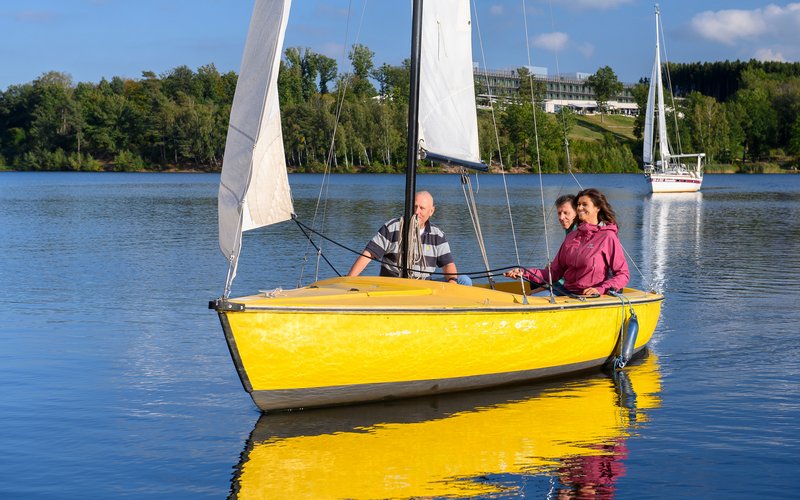 Ein gelbes Segelboot im Vordergrund mit einer Frau und zwei Männern als Insassen steuert schräg auf die Kamera zu. Die Frau, mit den Seilen des Segels in de Händen, und ein Mann sitzen am Heck des Bootes, während der andere Herr vorne im Boot Platz genommen hat. Hinter dem gelben Segelboot ist ein weiteres Segelboot in weißer Farbe auf dem Bostalsee zu erkennen.