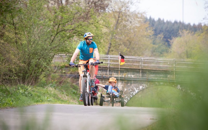 Ein Vater auf seinem Fahrrad fährt mit seinem Sohn, der sich in einem Liegerad befindet, über den flachen und asphaltierten Bahnradweg. Hinter ihnen folgt ein weiterer Mann auf seinem Rad.