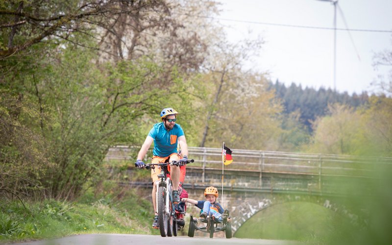 Ein Vater auf seinem Fahrrad fährt mit seinem Sohn, der sich in einem Liegerad befindet, über den flachen und asphaltierten Bahnradweg. Hinter ihnen folgt ein weiterer Mann auf seinem Rad.