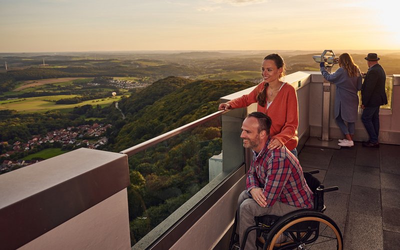 Ein Mann in einem Rollstuhl steht mit seiner Begleiterin auf dem Schaumbergturm und genießt von dort aus die Aussicht über das Sankt Wendeler Land, da das Geländer teilweise von Glas durchzogen ist.