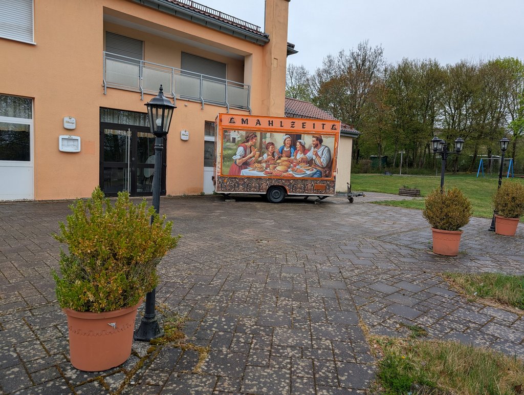 Frontalsicht auf einen geschlossenen Foodtruck, auf dem ein Gemälde abgebildet ist. Um den Foodtruck befinden sich Pflanzenkübel, die zu einer gemütlichen Atmosphäre beitragen. 