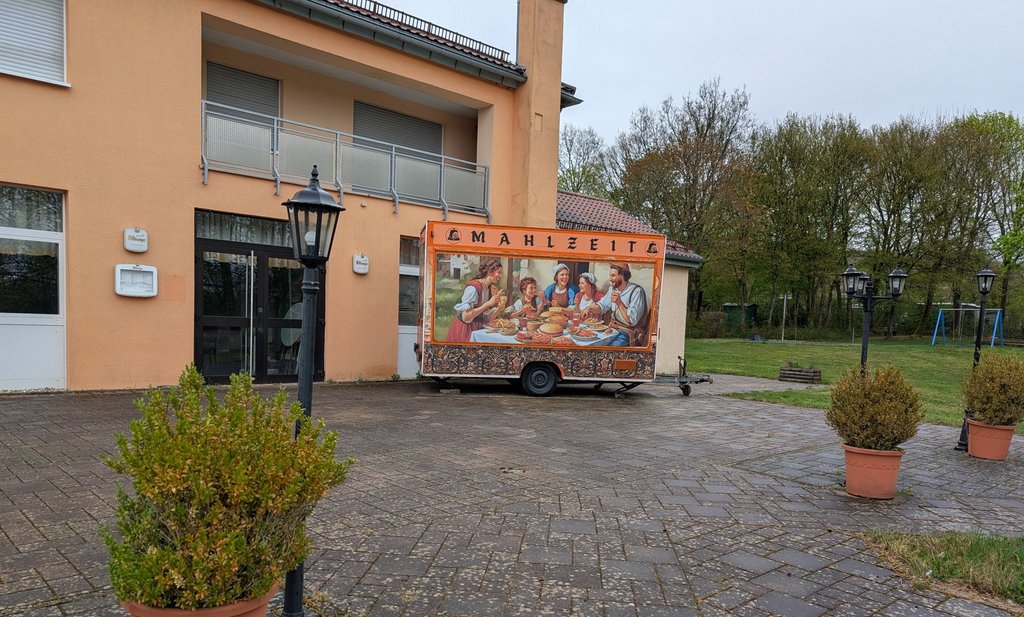 Foodtruck Mahlzeit Frontalsicht auf einen geschlossenen Foodtruck, auf dem ein Gemälde abgebildet ist. Um den Foodtruck befinden sich Pflanzenkübel, die zu einer gemütlichen Atmosphäre beitragen.