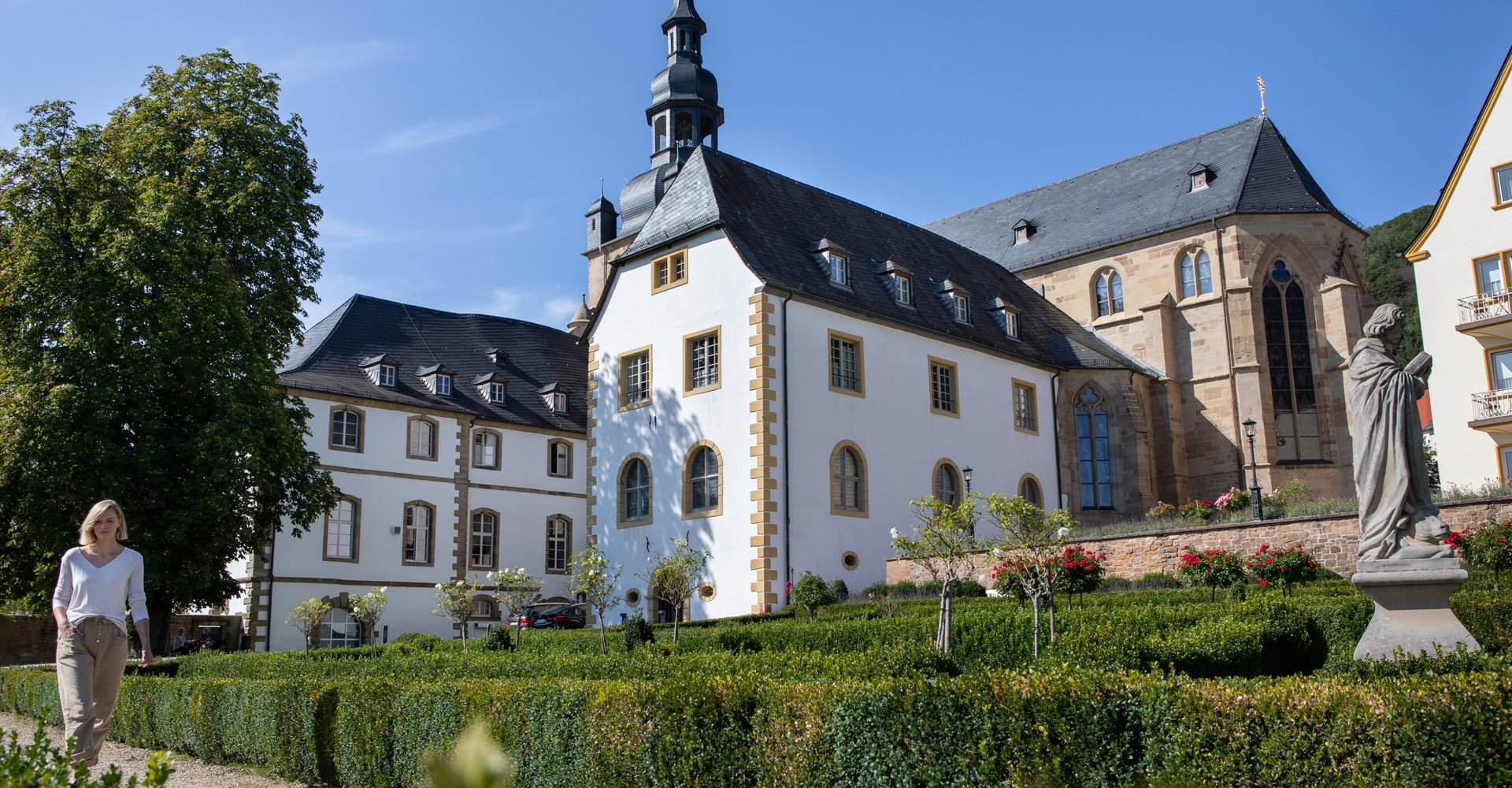 Eine Frau spaziert durch den Klostergarten der Abtei Tholey. Die Abtei sowie die Kirche sind im Hintergrund zu sehen.