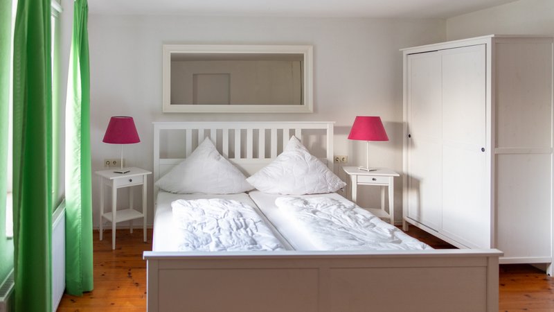 Ein Zimmer mit einem Doppelbett sowie zwei Nachttischen mit roten Nachttischlampen darauf. Vor dem Fenster hängen grüne Vorhänge und es gibt einen weißen Schrank.
