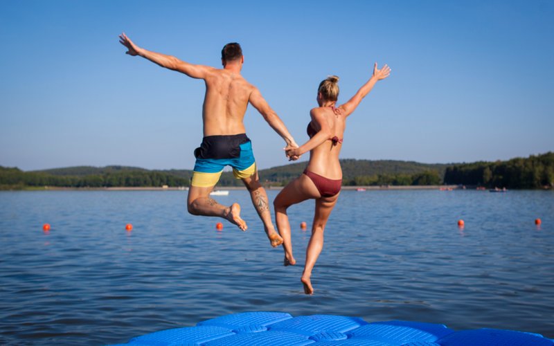 Ein junges Pärchen springt Hand in Hand im hohen Bogen von einer Schwimminsel aus in das blaue Wasser des Bostalsees.
