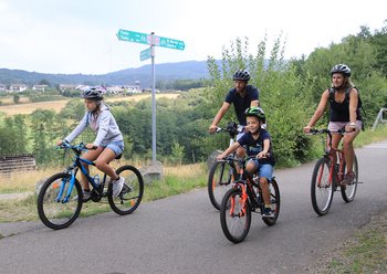 Eine Familie mit zwei Kindern fährt mit ihren Rädern auf dem asphaltierten Wendelinus-Radweg. Wegweiser zeigen ihnen den Weg. Die Strecke offenbart wunderbare Blicke auf das Sankt Wendeler Land.