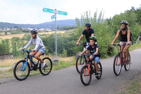 Eine Familie mit zwei Kindern fährt mit ihren Rädern auf dem asphaltierten Wendelinus-Radweg. Wegweiser zeigen ihnen den Weg. Die Strecke offenbart wunderbare Blicke auf das Sankt Wendeler Land.