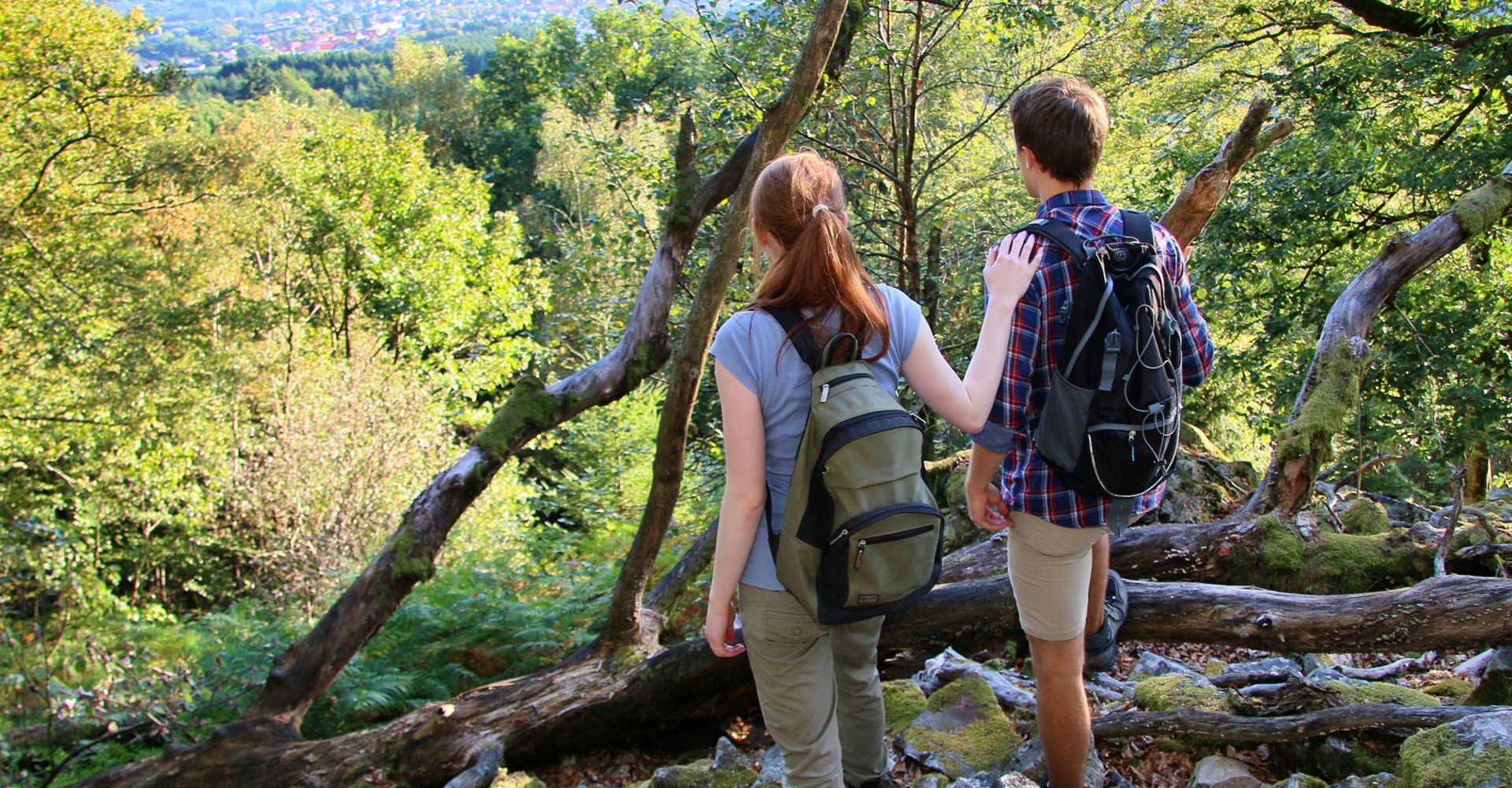 Ein junger Mann und eine junge Frau stehen in Wanderkleidung auf einem Hügel und schauen auf das grüne Tal mit seinen vielen Bäumen hinab.