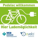 Logo Pedelec Willkommen Ein grün-weißes Schild, auf dem ein Fahrrad mit einem Ladekabel abgebildet ist. Darauf steht: "Pedelec willkommen. Hier Lademöglichkeiten". Zudem sind zwei Logos abgebildet.