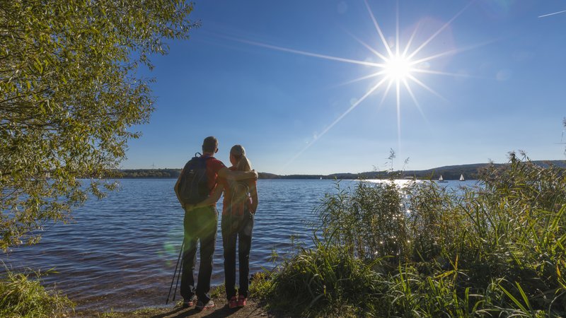 Ein Mann und eine Frau stehen, sich umarmend, am Ufer des Bostalsees und beobachten das Treiben auf dem Wasser. Die Sonne steht hoch am Himmel.