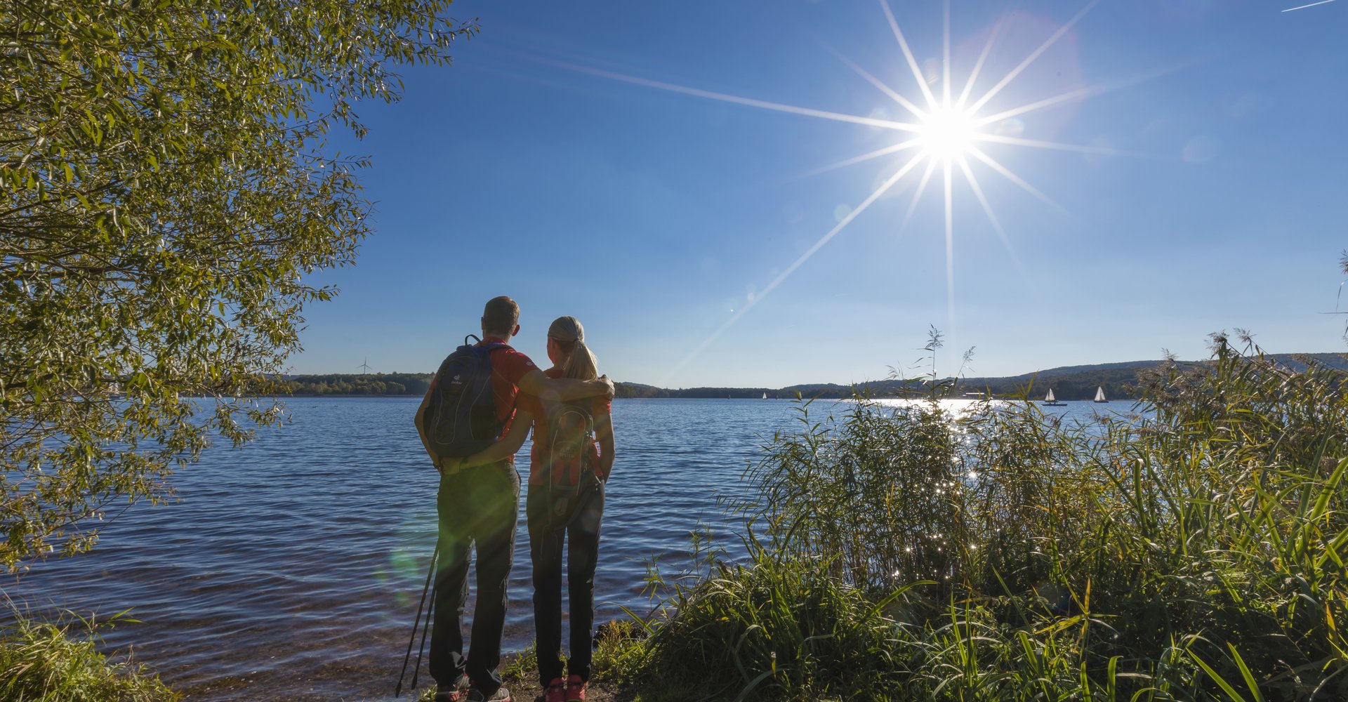 Ein Mann und eine Frau stehen, sich umarmend, am Ufer des Bostalsees und beobachten das Treiben auf dem Wasser. Die Sonne steht hoch am Himmel.