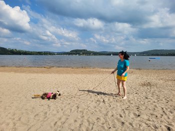 Eine Frau steht auf dem Sand des Strandes im Strandbad Bosen. Hinter ihr ist das Wasser des Bostalsees zu sehen. Neben ihr wälzt sich ein Hund im Sand.