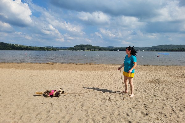 Eine Frau steht auf dem Sand des Strandes im Strandbad Bosen. Hinter ihr ist das Wasser des Bostalsees zu sehen. Neben ihr wälzt sich ein Hund im Sand.