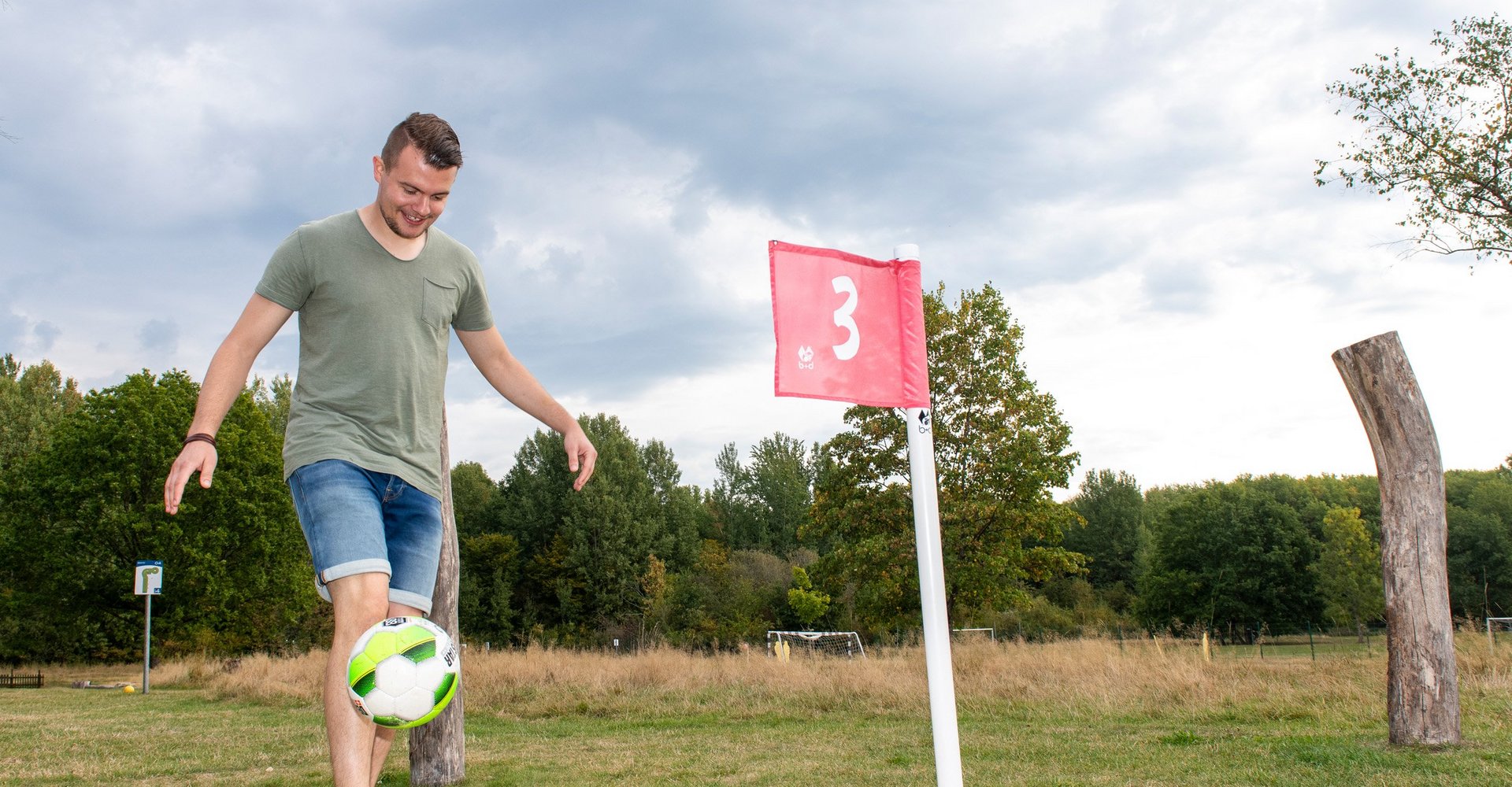 Ein Mann in kurzer Freizeitkleidung versucht einen Fußball in ein großes Golfloch einzulochen. Dieses ist mit einer roten Fahne gekennzeichnet.