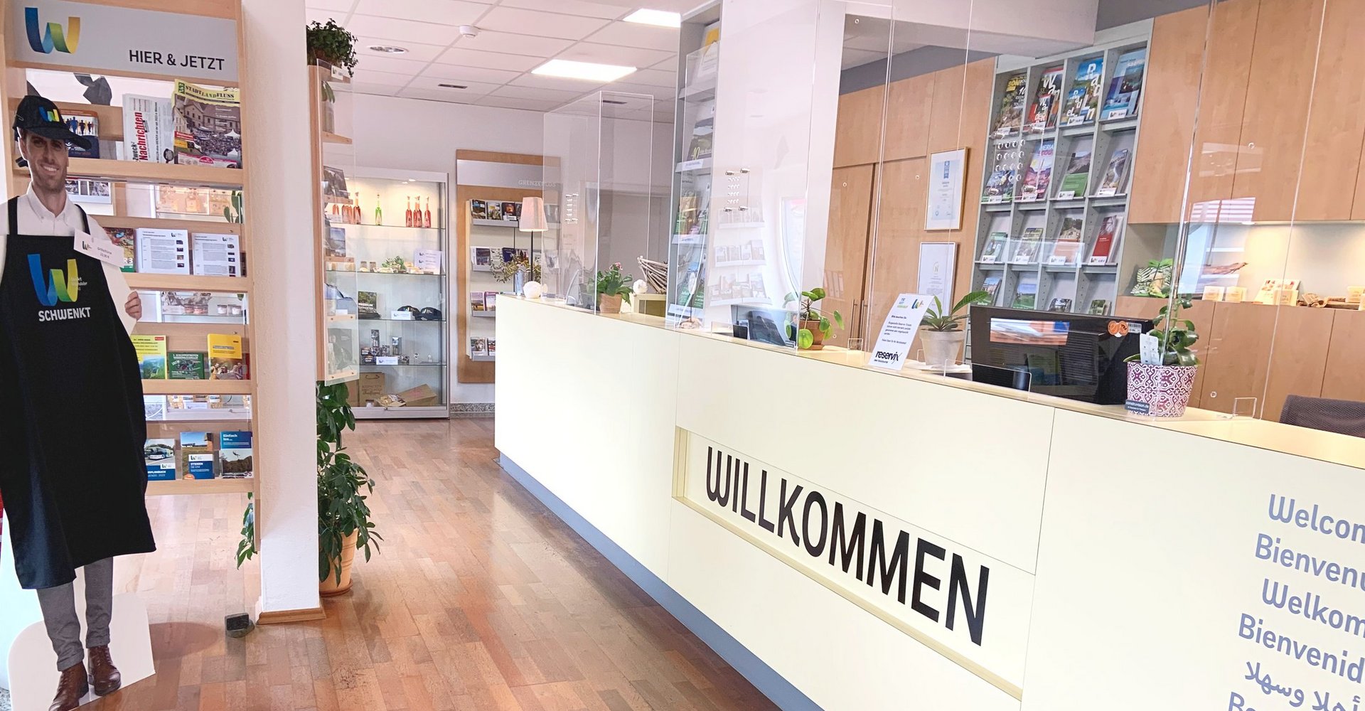 Der Eingangsbereich der Tourist-Information begrüßt die Gäste mit einem Willkommen an der langen Theke. Rundherum befinden sich Informationsbroschüren und Verkaufsartikel