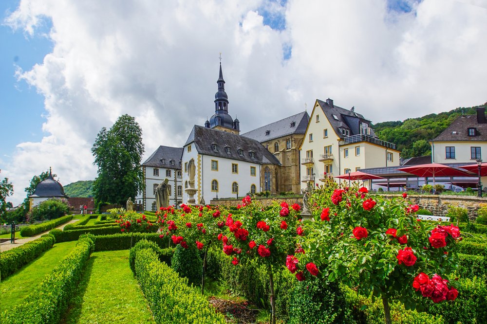 Individuelle Gruppenführung Abtei_Gemeinde Tholey Die Abtei Tholey steht im Zentrum des Bildes und wird von außen gezeigt. Davor erstreckt sich der Klostergarten, in dem rote Rosen blühen und das Gras saftig grün erscheint.