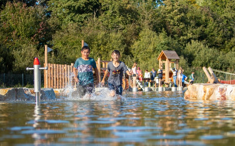 Zwei Jungen laufen durch das Wasser auf dem Wasserspielplatz während die anderen Kinder an den Spielstationen rutschen, klettern und 