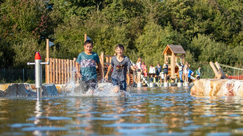 Zwei Jungen laufen durch das Wasser auf dem Wasserspielplatz während die anderen Kinder an den Spielstationen rutschen, klettern und 
