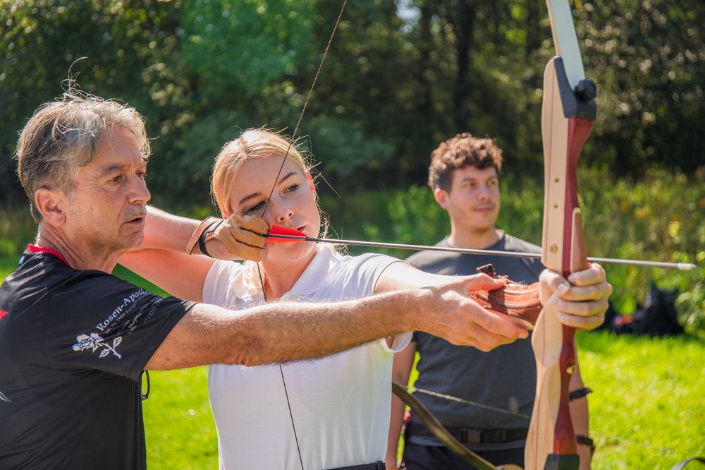 Eine Frau wird von einem Trainer im Bogenschießen angelernt. Sie ist mit Pfeil  und Bogen ausgestattet. Im Hintergrund ist eine weitere Person zu sehen, die der Erklärung lauscht.
