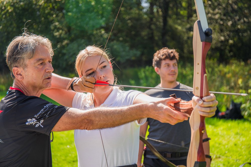 Eine Frau wird on einem Trainer im Bogenschießen angelernt. Sie ist mit Pfeil und Bogen ausgestattet. Im Hintergrund ist eine weitere Person zu sehen, die der Erklärung lauscht.