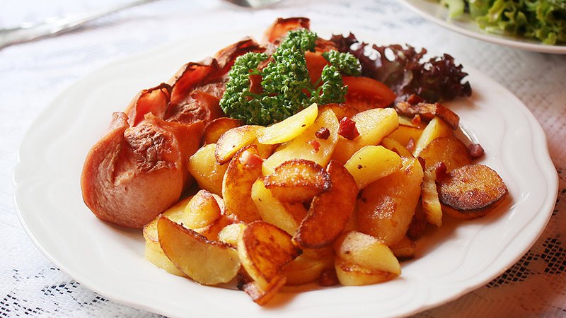 Auf dem Teller wird das saarländisches Gericht Lyonerpfanne serviert. Es besteht aus gebratenem Lyoner und Bratkartoffeln mit Schinkenwürfeln. Im Hintergrund steht ein Teller mit Salatbeilage.