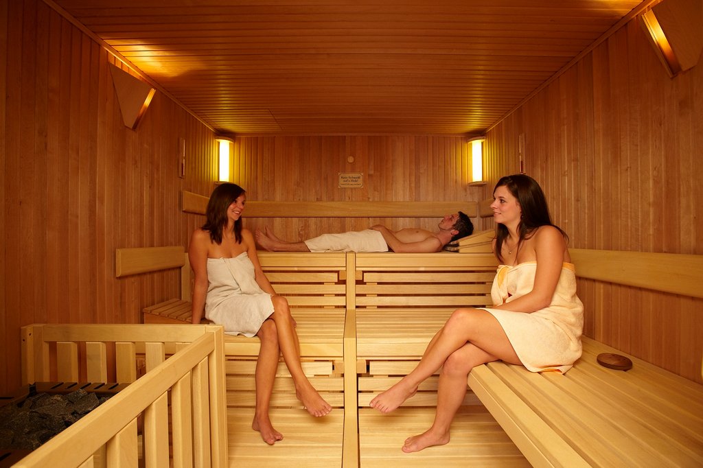 2 Frauen sitzen sich, mit Handtüchern bekleidet, in einer Sauna gegenüber, während ein Mann, ebenfalls mit Handtuch bedeckt, dahinter auf einer der Bänke liegt.