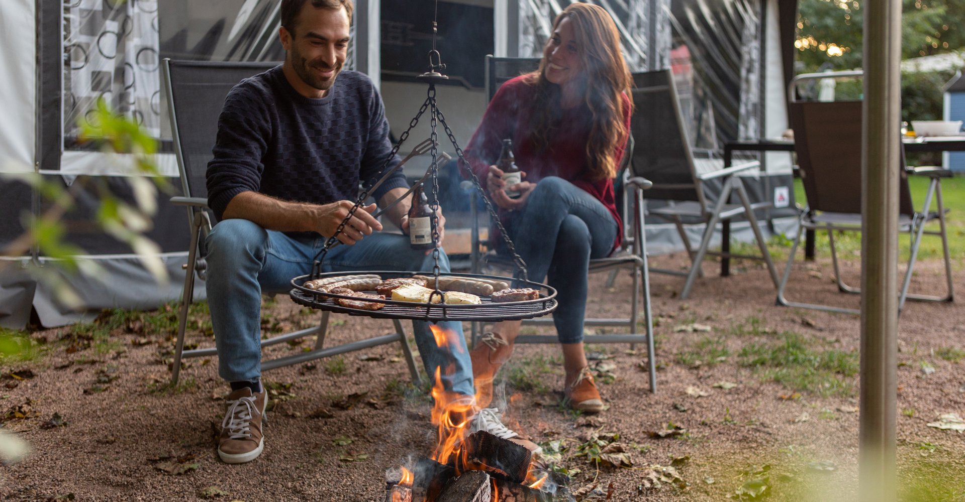 Eine Frau und ein Mann sitzen vor einem Zelt am Feuer und grillen beziehungsweise Schwenken ihr Grillgut.
