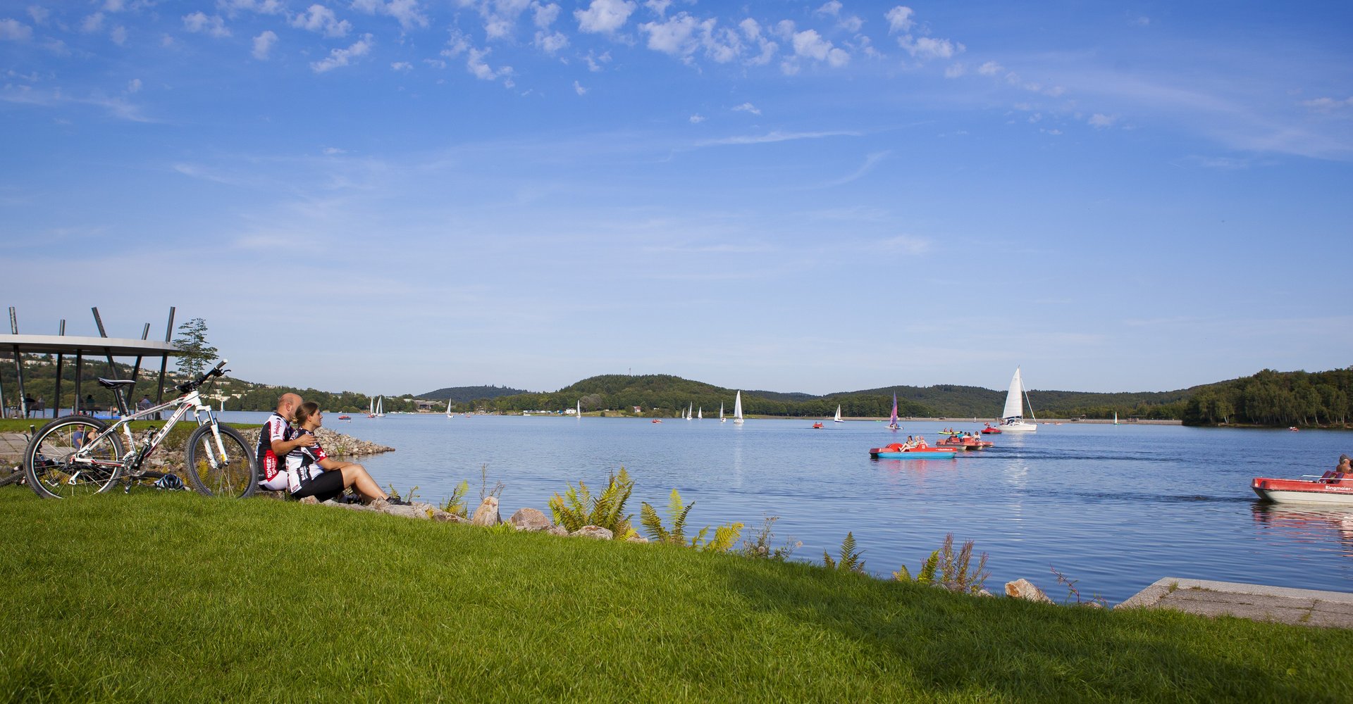 2 Radfahren sitzen am Ufer des Bostalsees und schauen den Tretbootfahrern und Seglern auf dem Wasser zu.