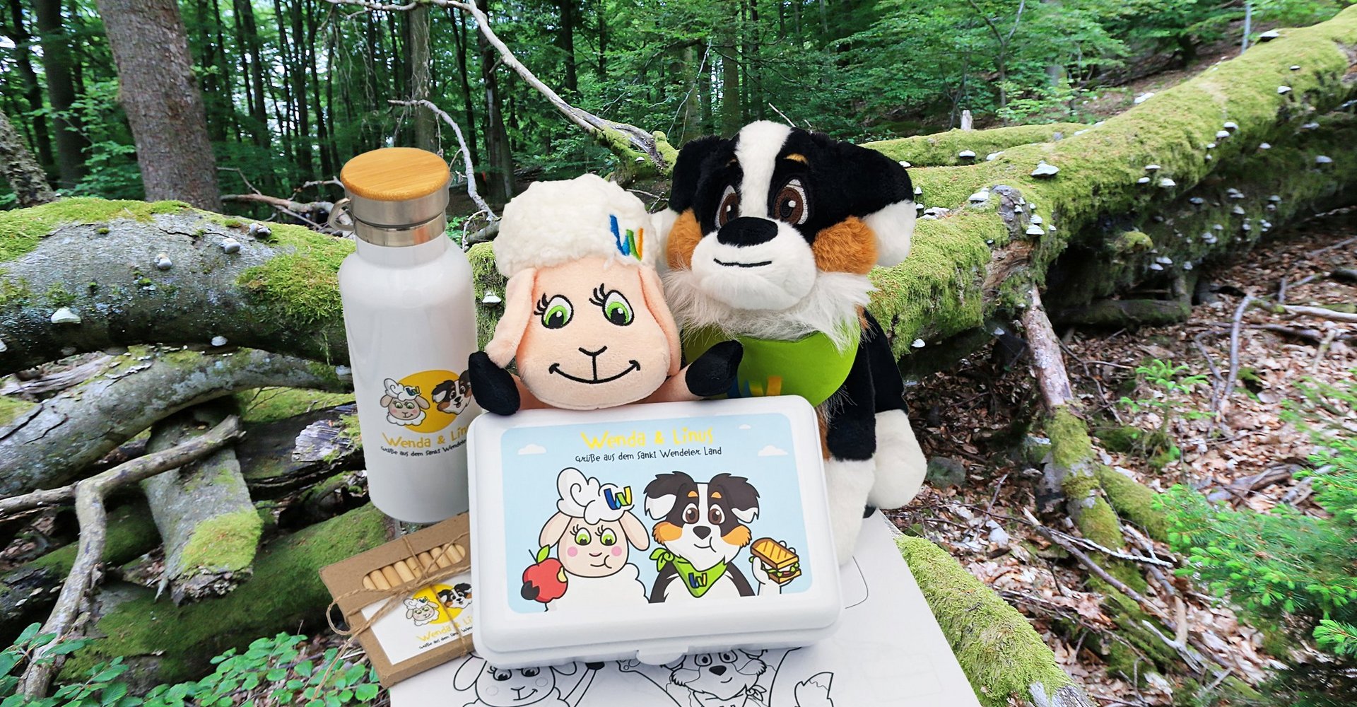 Die Plüschtiere Wenda, das Schaf, und Linus, der Hund,, mit einer Trinkflasche, einer Brotdose und Malstiften sowie einem Malbuch mit dem Wald im Hintergrund.