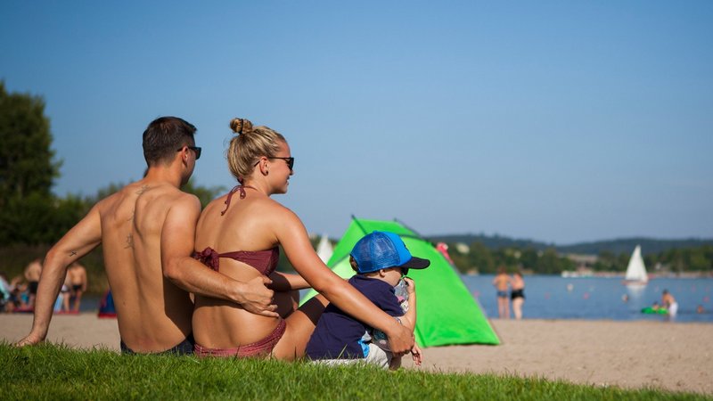 Ein junges Paar mit kleinem Kind sitzt auf der Liegewiese eines Stranbads am Bostalsee und schaut auf den See. Im Hintergrund ist eine grüne Strandmuschel zu sehen.