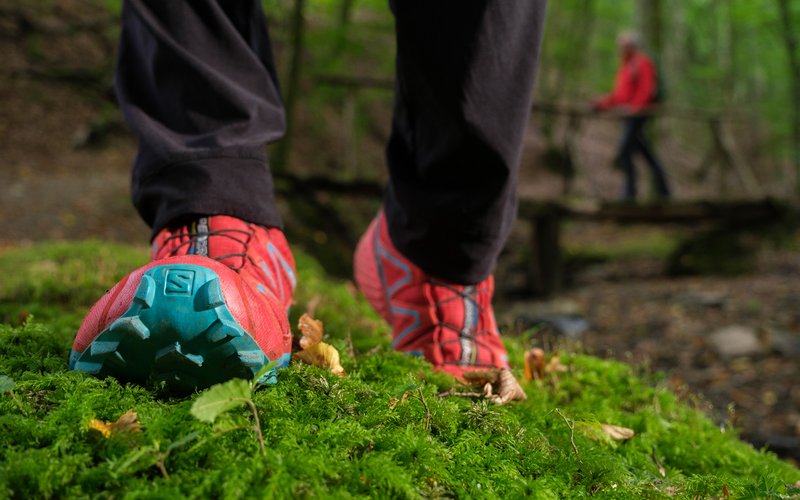 In einer Nahaufnahme sind rote Wanderschuh mit blauer Sohle, die auf grünen Waldboden auftreten, zu sehen. Im Hintergund ist ein weiterer Wanderer, leicht verschwommen, zu sehen.