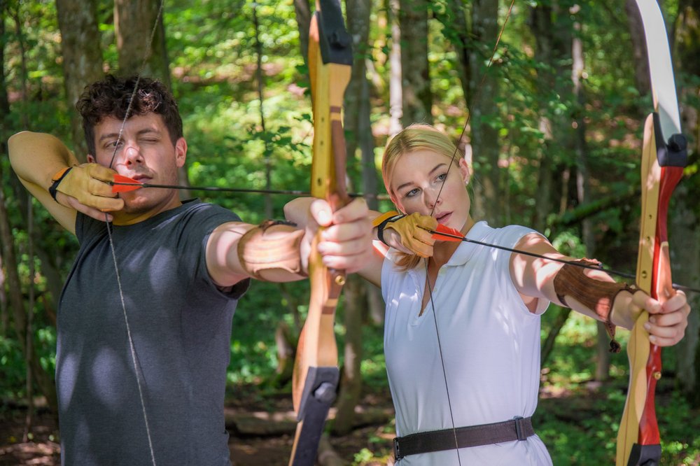 Zwei Personen stehen mit Pfeil und Bogen nebeneinander im Wald und visieren ihr Ziel an.