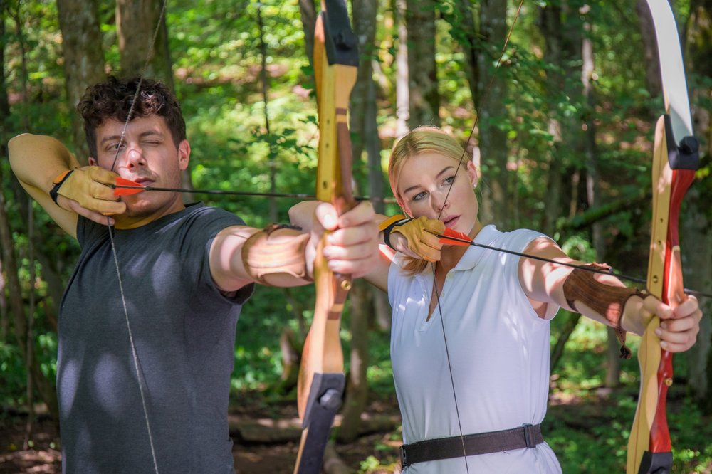 Gruppenkurs Bogenschießen Zwei Personen stehen mit Pfeil und Bogen nebeneinander im Wald und visieren ihr Ziel an.