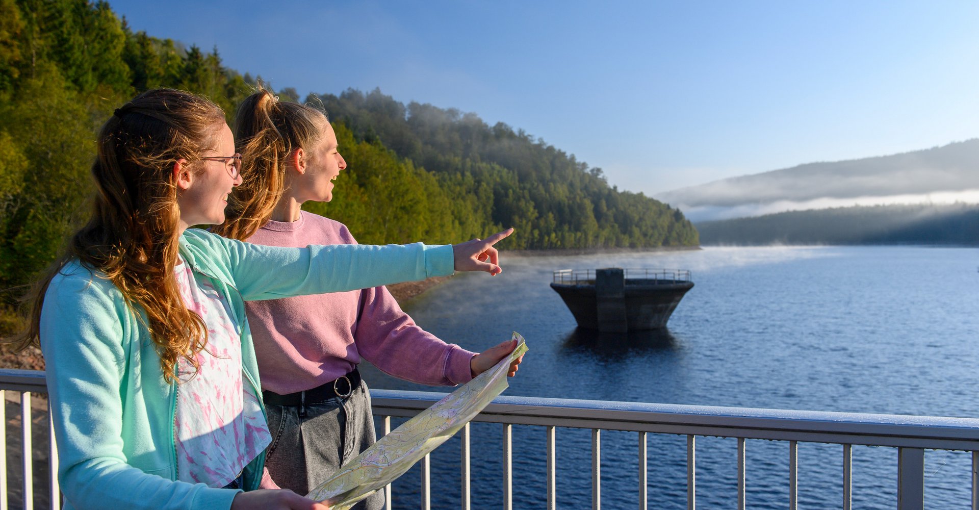 Zwei Frauen stehen auf der Staumauer der Talsperre. Eine Frau hat eine Karte in der Hand und zeigt auf etwas in der Ferne.