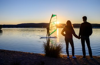 bei Sonnenuntergang auf dem Bostalsee steht ein Windsurfer auf seinem Bord und schaut zum Ufer, wo gleichzeitig ein junges Pärchen Hand in Hand steht.