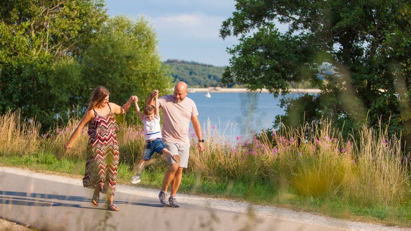 Ein Paar mit einem kleinem Jungen geht auf dem Seerundweg spazieren. Sie spielen mit dem Jungen das Spiel "Engelchen flieg". Er wird von beiden an seinen Armen in die Luft gehoben. Im Hintergrund ist ein Blick auf den See zu erhaschen.