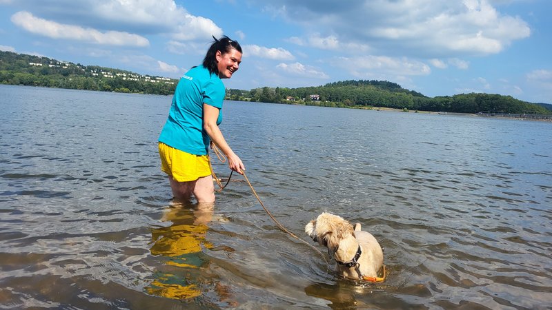 Eine Frau steht mit ihrem kleinen beigefarbenen Hund im Wasser an der Hundebadestelle an der Staumauer