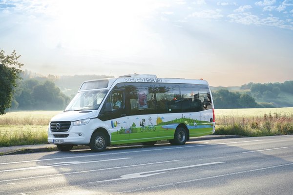 Ein weißer Bus mit einer bunten Beklebung und der Aufschrift "Steig ein fahr mit" fährt über eine Landstraße.
