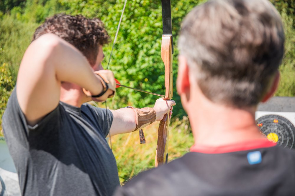 In einer Nahaufnahme ist im Vordergrund ein Mann mit Pfeil und Bogen zu sehen. Er bekommt Anweisungen von seinem Trainer, der hinter ihm steht.