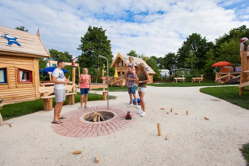 Es ist der zentrale Grillplatz des Märchendorfs mit den Märchenhäusern auf dem Campingplatz Bostalsee zu sehen. Ein ppar Campinggäste haben sich um das Schwenkergerüst versammelt und nehmen ein Erfrischungsgetränk zu sich.