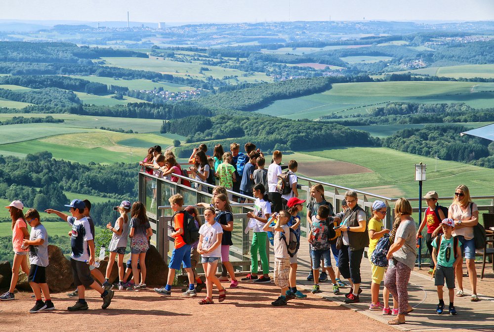 Gruppenangebote 4 Varianten Gemeinde Tholey Eine Schulklasse mit Lehrerin tummelt sich auf der Aussichtsplattform des Schaumbergplateaus und genießt die Aussicht von oben über die grüne, hügelige Landschaft.